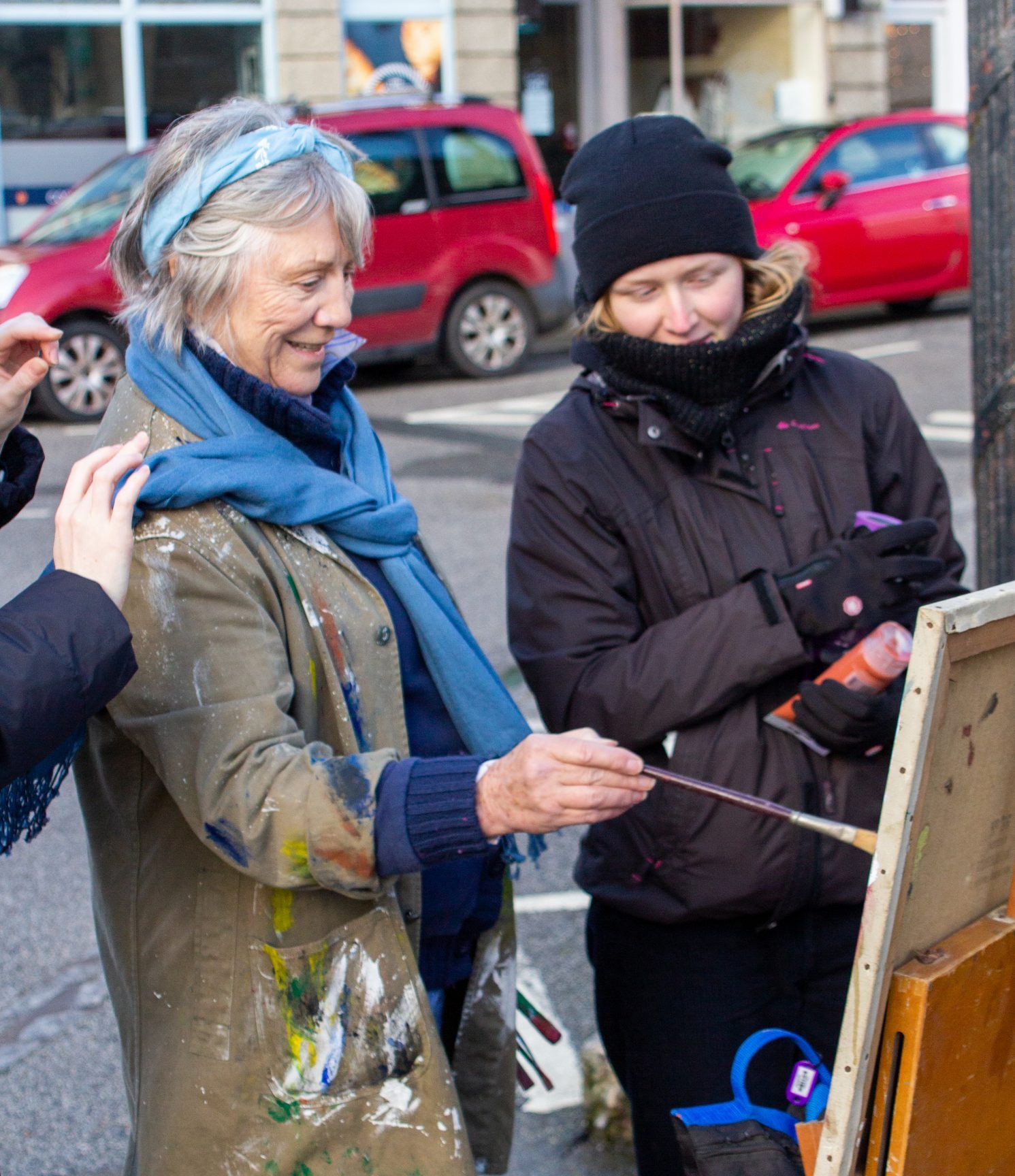 two women standing in front of a canvas. One is painting and the other is holding paints.